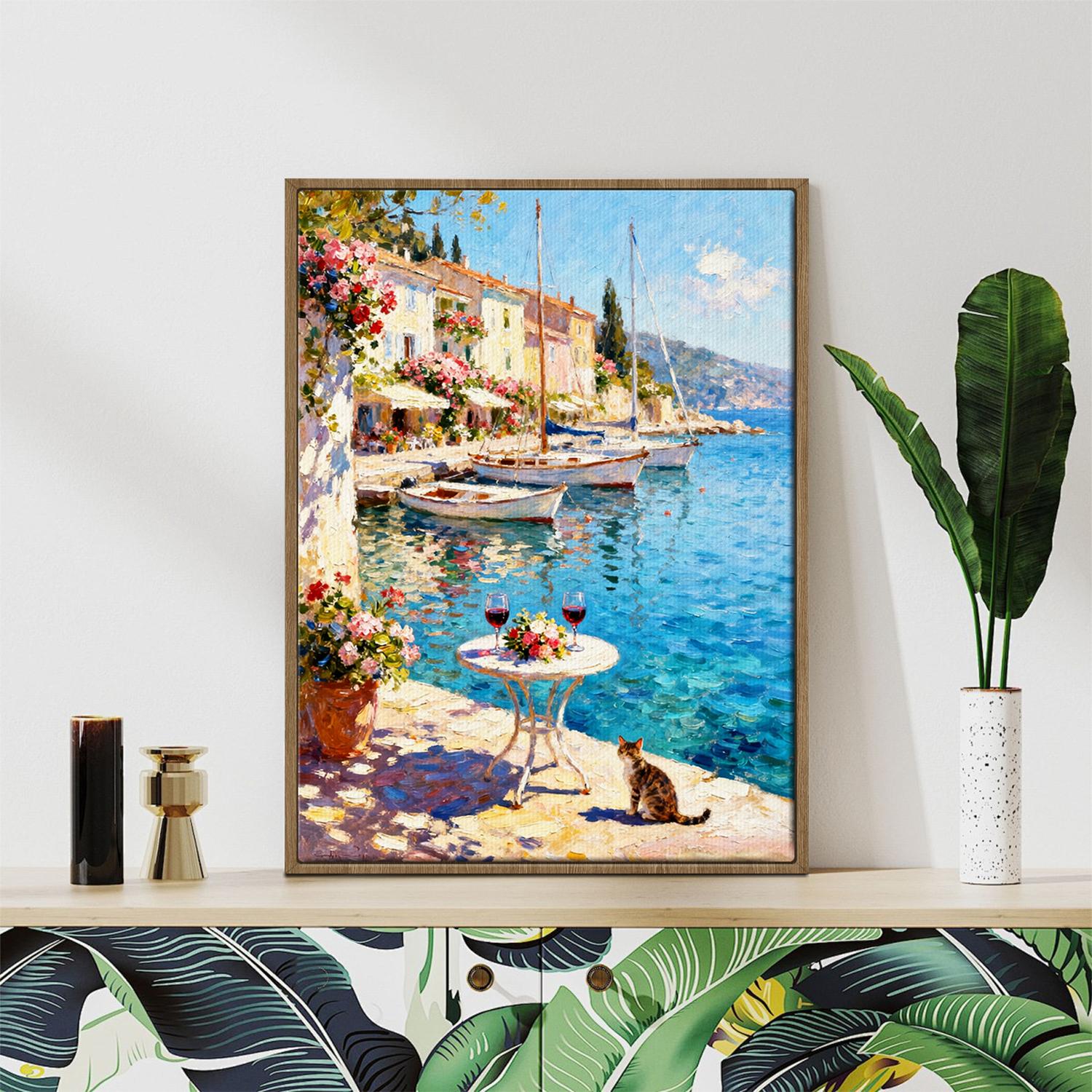 light oak color frame wall art canvas painting showing a sunny seaside town with a table of red wine and flowers on the pier and a cat.